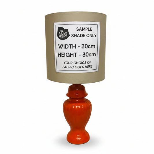 A table lamp sample with a beige fabric shade, a glossy orange-red ceramic base, and a white paper label that reads 'SAMPLE SHADE ONLY' with dimensions and fabric placeholder text, photographed against an off-white wall.