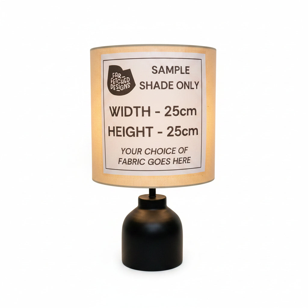 A table lamp with a black cylindrical base and a beige shade, featuring a white paper label that reads 'SAMPLE SHADE ONLY' with dimensions 'WIDTH - 25cm' and 'HEIGHT - 25cm'.