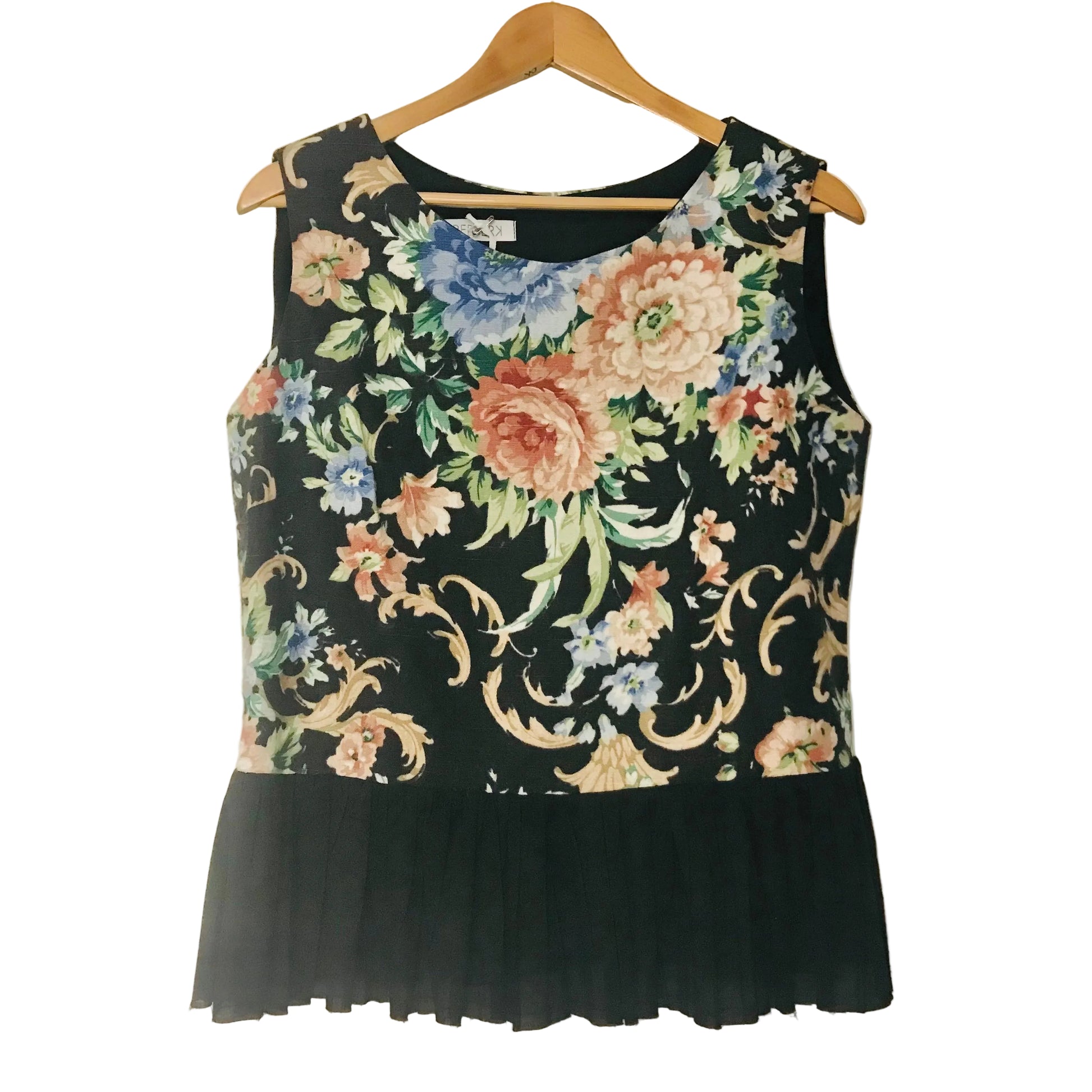 A sleeveless black top with a colorful floral and scroll pattern on the upper bodice and a solid black ruffled hem, hanging on a wooden hanger against a plain off-white wall.
