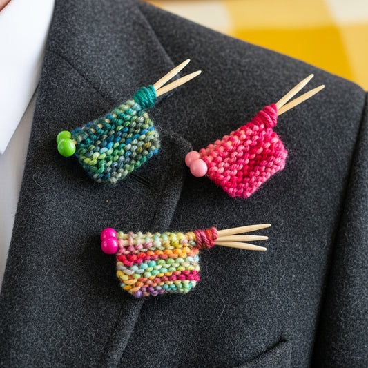 Three small knitted yarn pieces on wooden double-point needles, each attached to a white card labeled 'Knitter's Brooch,' arranged on a yellow textured blanket.