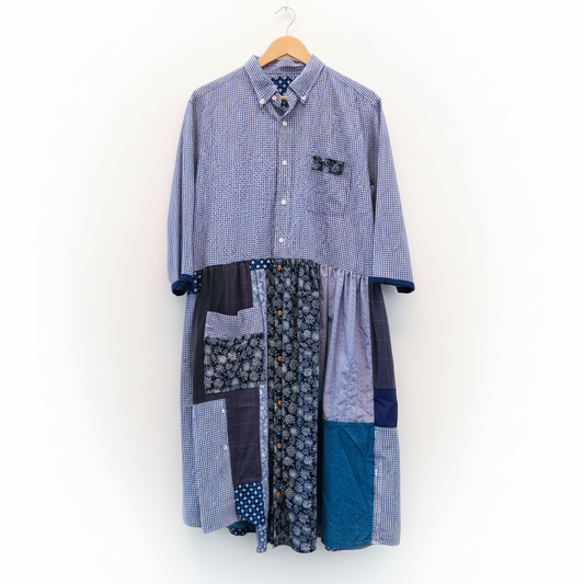 A patchwork long-sleeve button-up shirt dress hanging on a wooden hanger against a plain white wall. The dress features multiple fabric panels in blue, navy, and gray patterns, including gingham, floral, and solid textures.