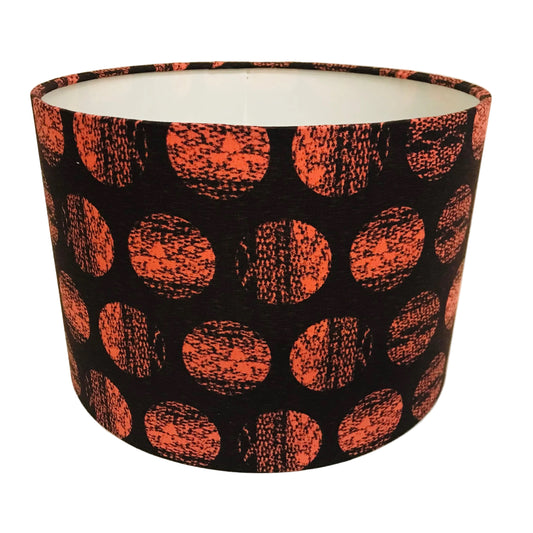 A cylindrical lampshade with a black background and a pattern of large orange-red textured polka dots, photographed against a plain off-white background.