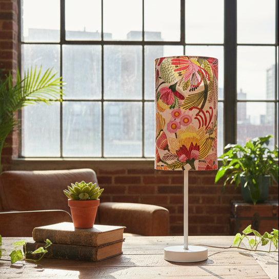 Floral-patterned lampshade on a table with books and a plant in a room with large windows.