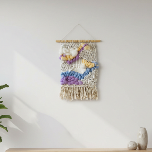 Woven wall art with colorful yarn on a white wall, next to a plant and small vase.
