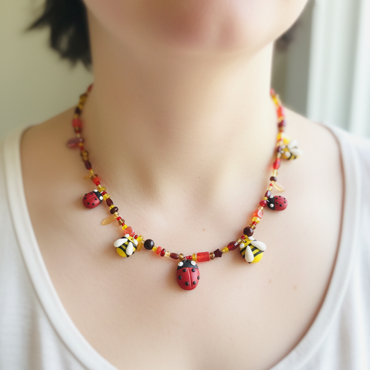 A close-up of a person wearing a colorful beaded necklace with ladybug and bumblebee charms. The necklace features red, yellow, black, and white beads, with a prominent red ladybug charm at the center, worn over a white tank top.