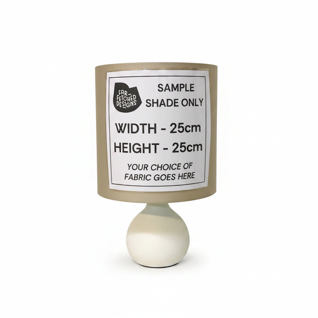 A cylindrical fabric lampshade with a white printed sample label reading 'SAMPLE SHADE ONLY' and dimensions, mounted on a white round table against a plain off-white wall.