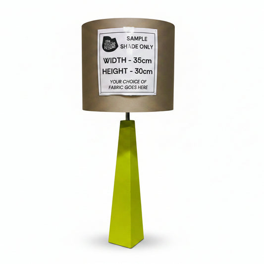 A custom fabric table lamp shade in a metallic taupe-brown color, mounted on a bright lime green geometric pedestal base. A white paper label is pinned to the front of the shade with dimensions and customization details.