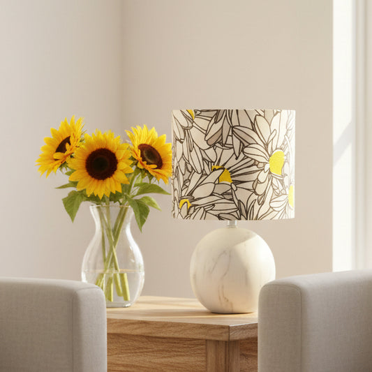 A table lamp with a white marble base and a drum shade featuring a stylized daisy pattern in gray, white, black, and yellow.