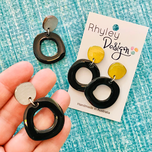 Black and yellow earrings held by a hand with 'Rhyley Design' packaging on a blue textured background.
