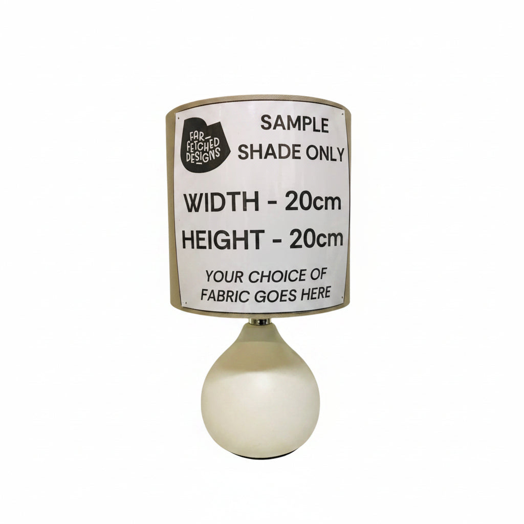 A table lamp with a white ceramic base and a custom fabric drum shade, featuring a printed sample label that reads 'SAMPLE SHADE ONLY' and dimensions 'WIDTH - 20cm HEIGHT - 20cm'.