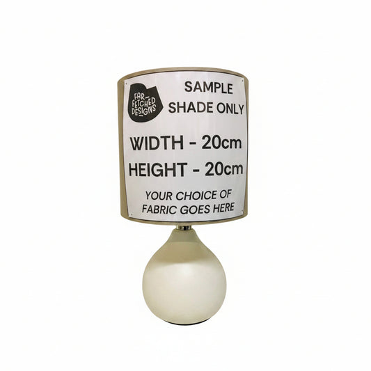 A table lamp with a white ceramic base and a custom fabric drum shade, featuring a printed sample label that reads 'SAMPLE SHADE ONLY' and dimensions 'WIDTH - 20cm HEIGHT - 20cm'.