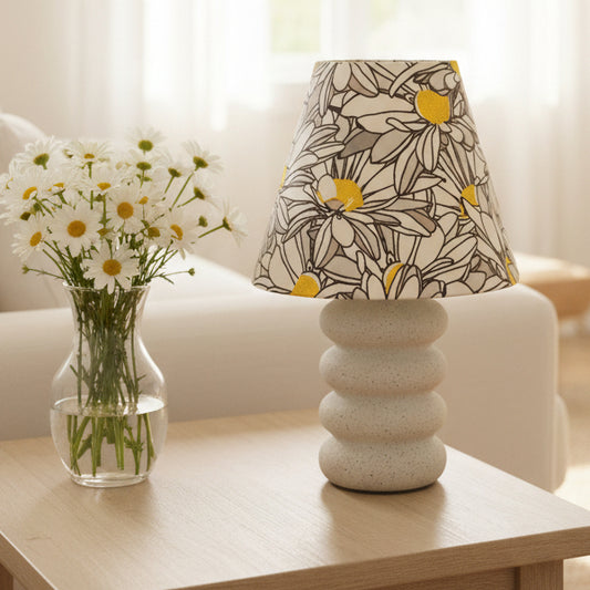 A table lamp with a white fabric shade featuring a black and gray daisy pattern with bright yellow centers, and a textured, tiered off-white ceramic base.