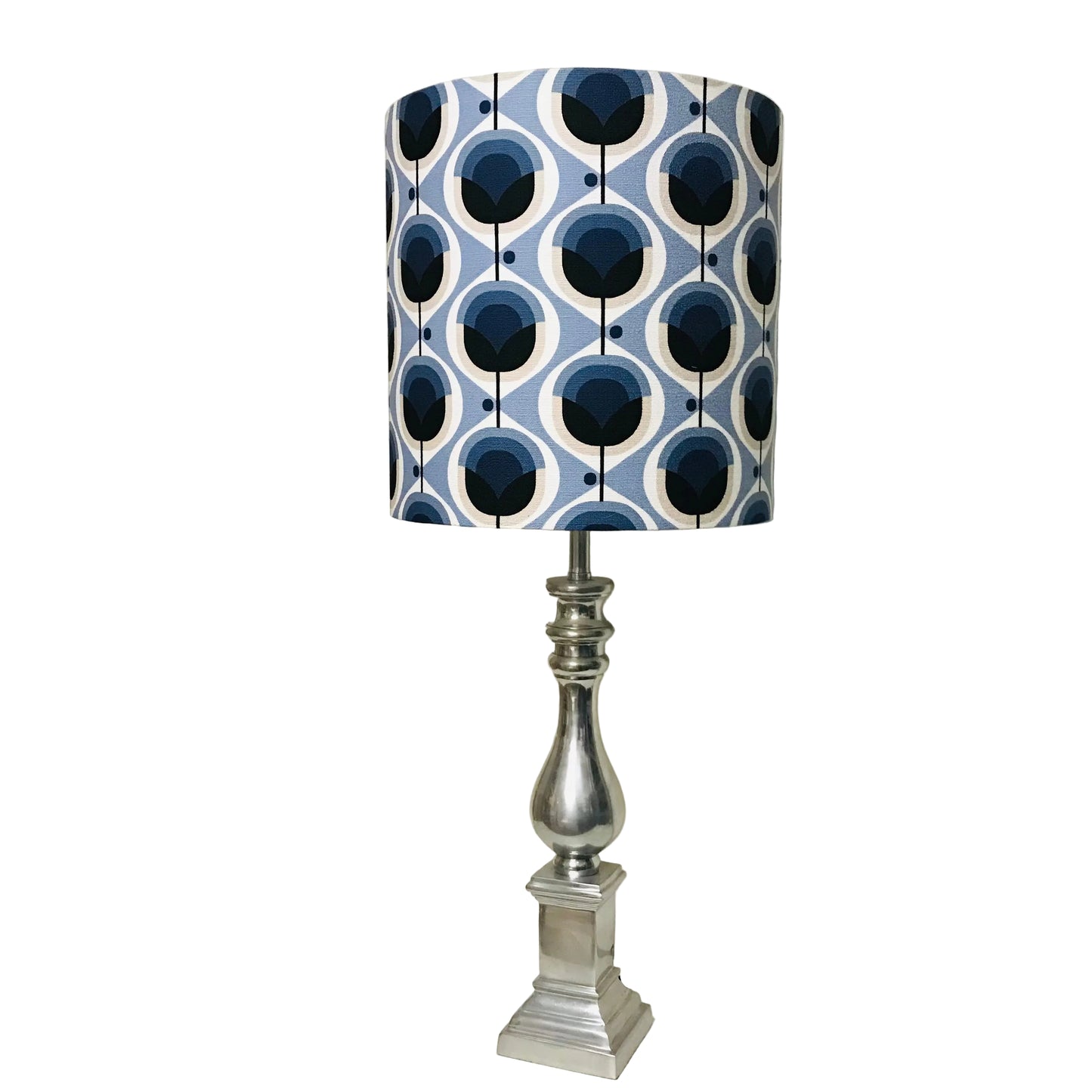 A table lamp with a cylindrical drum shade featuring a blue, white, and navy geometric pattern, mounted on a polished silver metal base and set on a white tabletop.