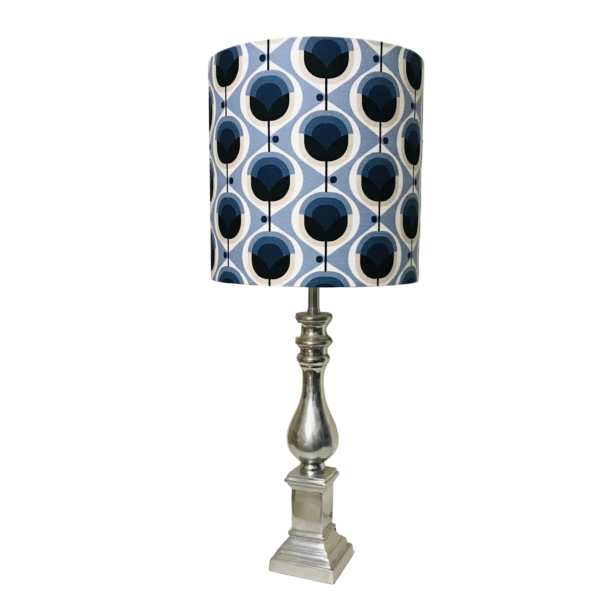 A table lamp with a cylindrical drum shade featuring a blue, white, and navy geometric pattern, mounted on a polished silver metal base and set on a white tabletop.