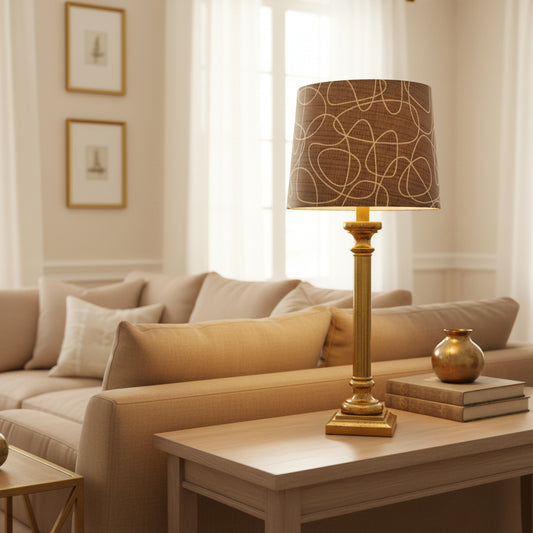 A table lamp with a square gold-colored metallic base and a cylindrical drum shade featuring a dark gray and beige abstract line pattern