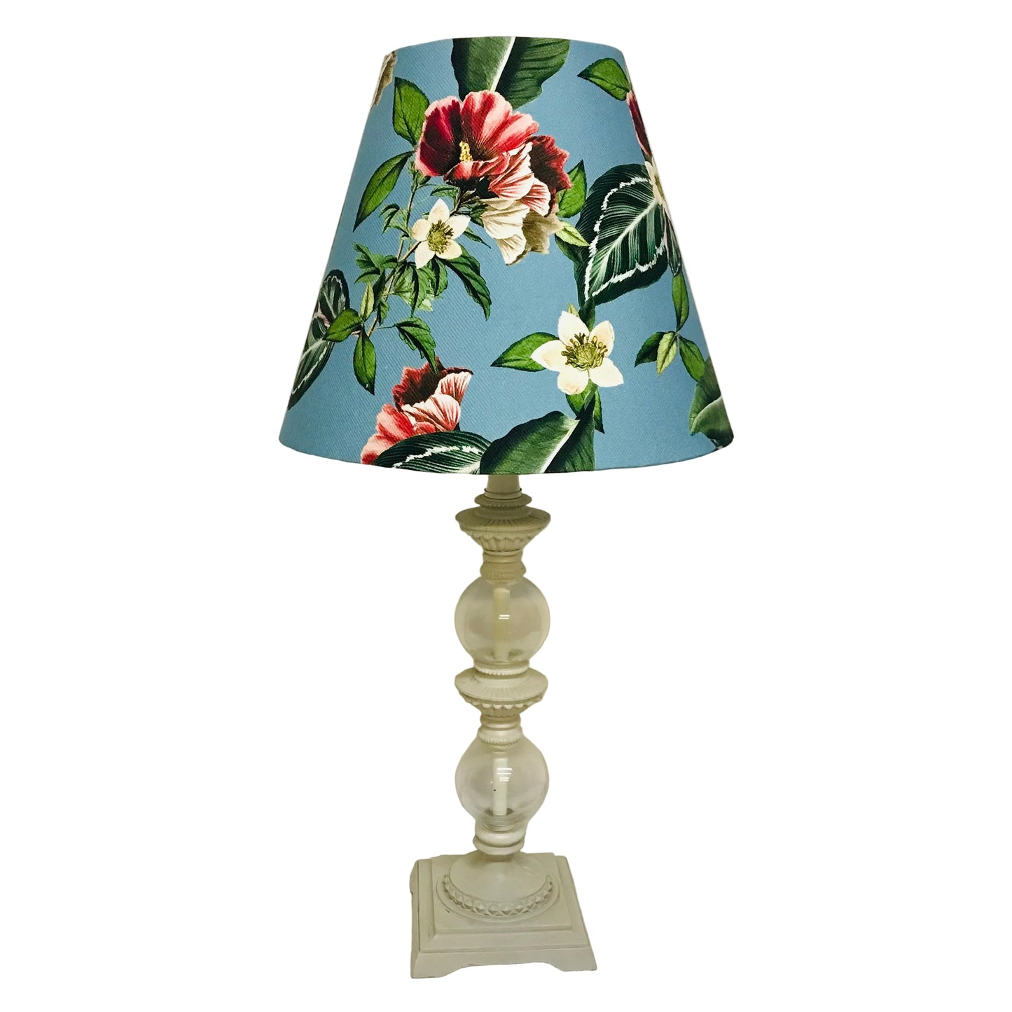 A table lamp with a blue floral fabric shade and a cream-colored glass and metal base, set against a plain off-white wall.