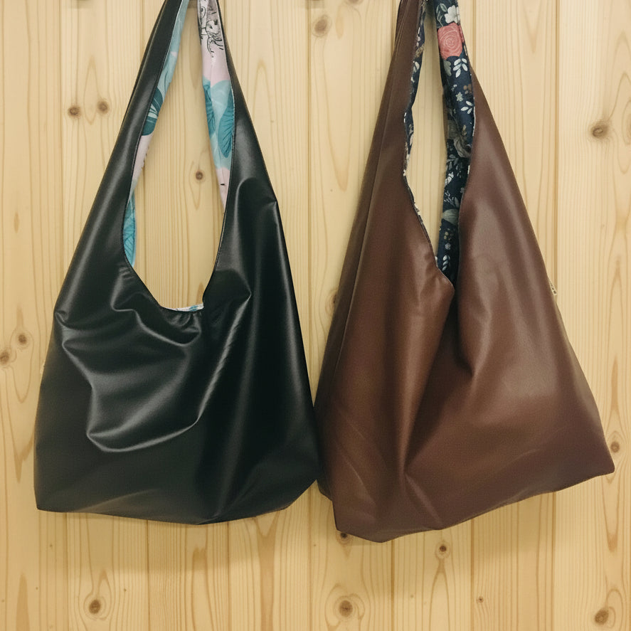 Two slouchy shoulder bags hanging from wall hooks on a white wall. The left bag is black faux leather with a pastel floral patterned strap lining, and the right bag is brown faux leather with a dark floral patterned strap lining.