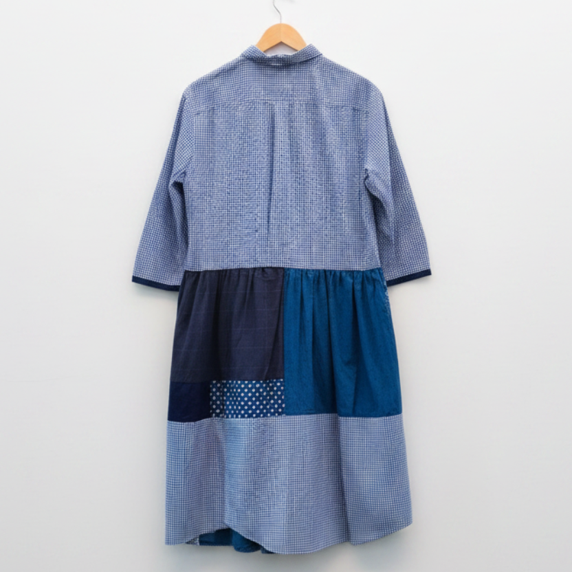 A patchwork blue shirt dress hanging on a wooden hanger against a white painted brick wall. The dress features a blue and white gingham pattern on the upper bodice and sleeves, a solid dark blue waistband, and various patchwork panels in blue, navy, and light blue gingham on the skirt.