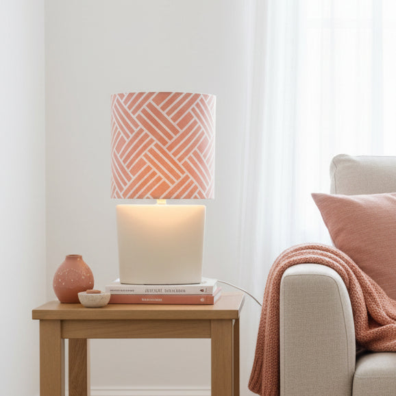 A table lamp with a cylindrical cream-colored base and a patterned shade featuring a terracotta pink and white geometric herringbone design