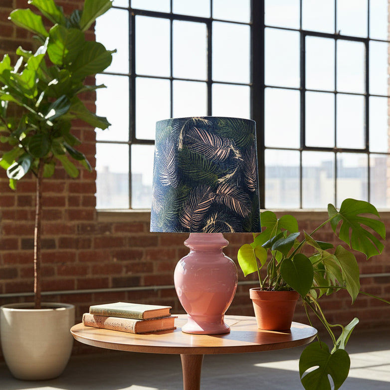 Pink lamp with a leaf-patterned shade on a wooden table, surrounded by plants in a room with large windows.