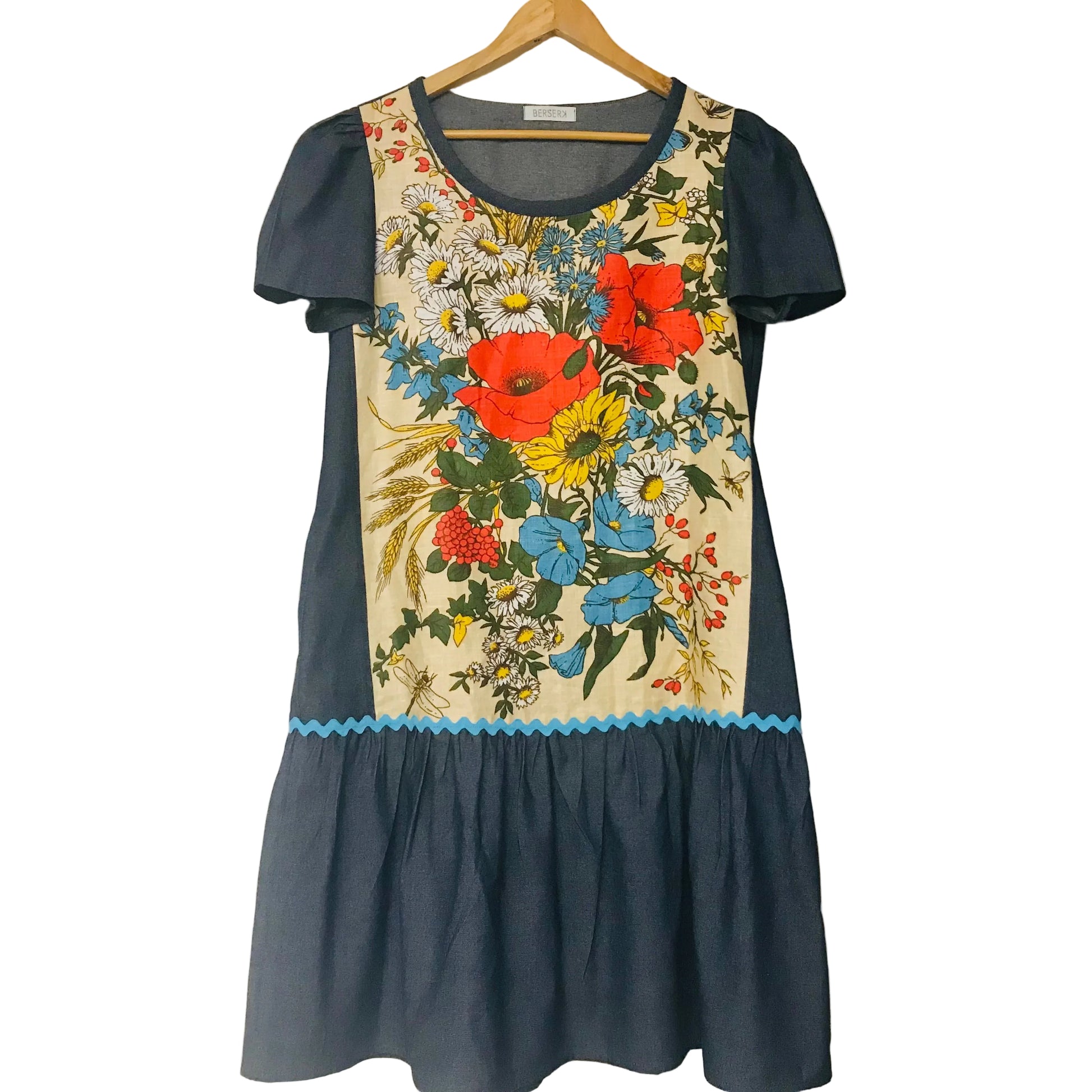 A short-sleeve dress hanging on a wooden hanger against a plain off-white wall. The dress has a dark navy blue base with a large, colorful floral print on the front panel and a light blue scalloped trim at the waist.