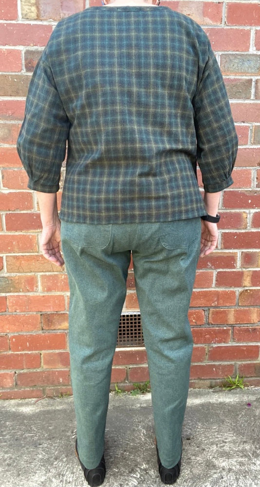 Person wearing a plaid shirt and green pants standing against a brick wall.