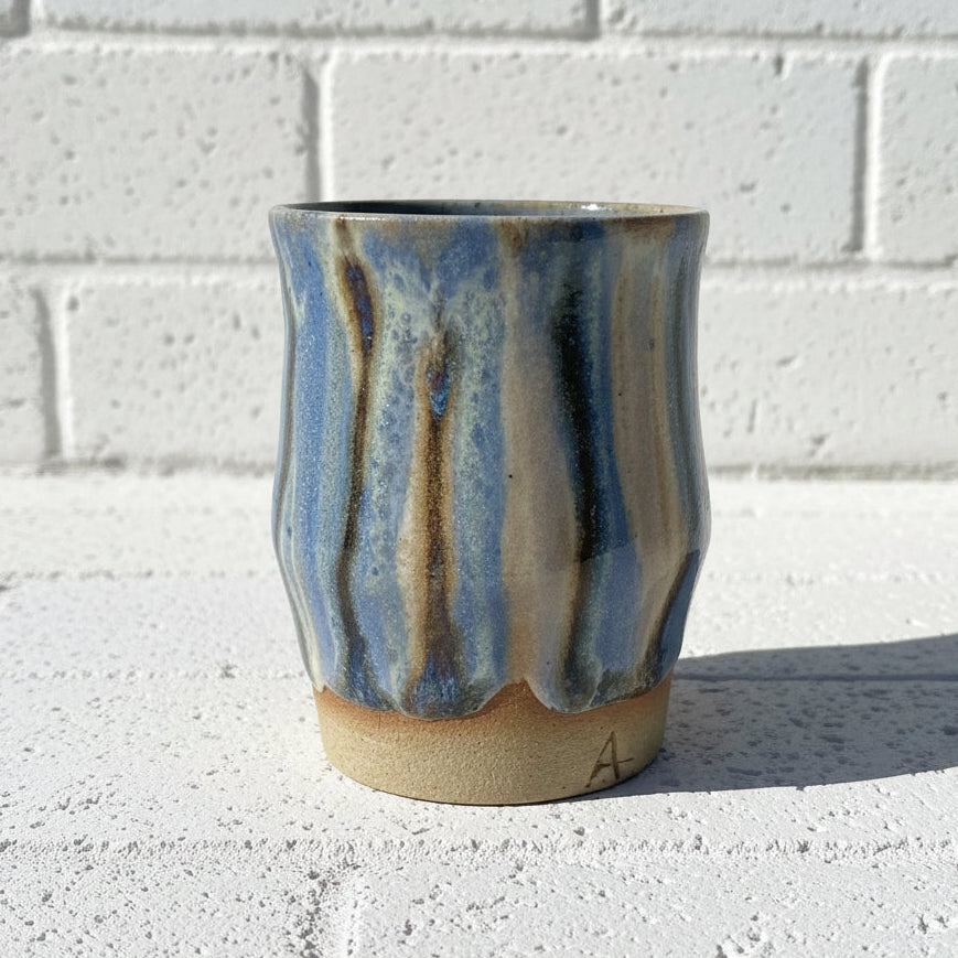 Handmade ceramic cup with blue and brown glaze on a concrete surface with a white brick wall background.