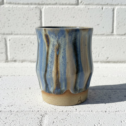 Handmade ceramic cup with blue and brown glaze on a concrete surface with a white brick wall background.