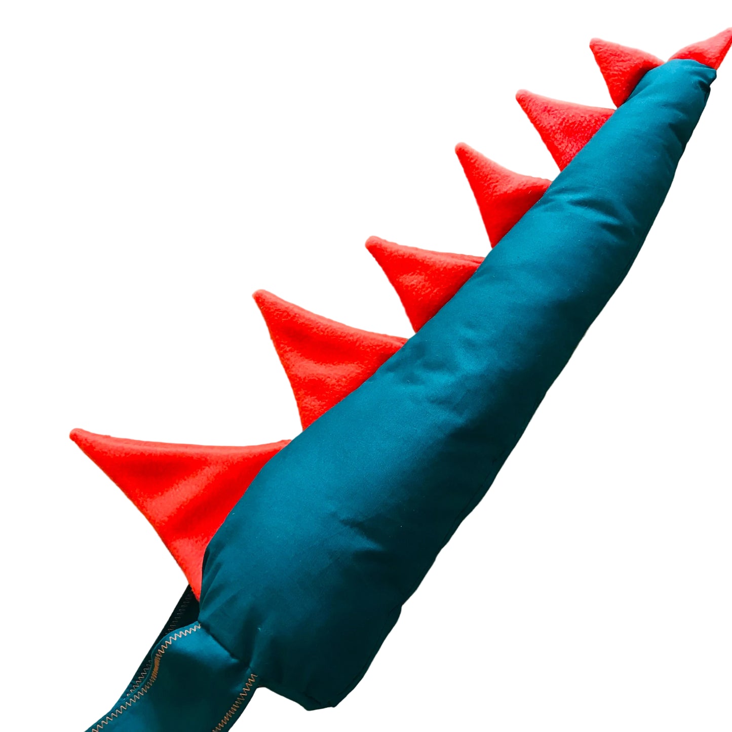 A teal and red dinosaur tail toy or pillow, featuring a teal fabric body and bright red felt spikes, laid diagonally on a light wood background.