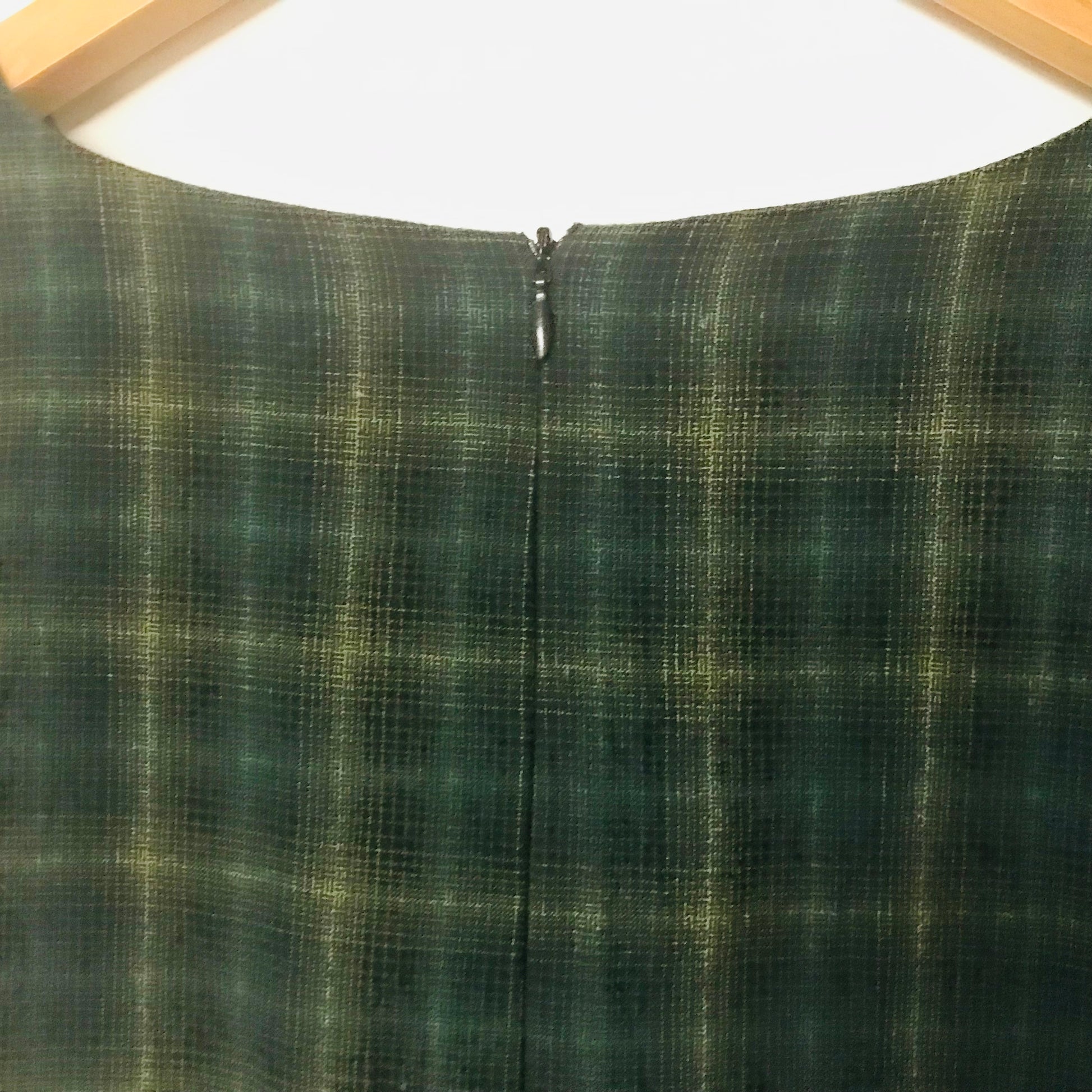 Close-up of the back of a dark green plaid garment with a central zipper, hanging on a wooden hanger against a white background.