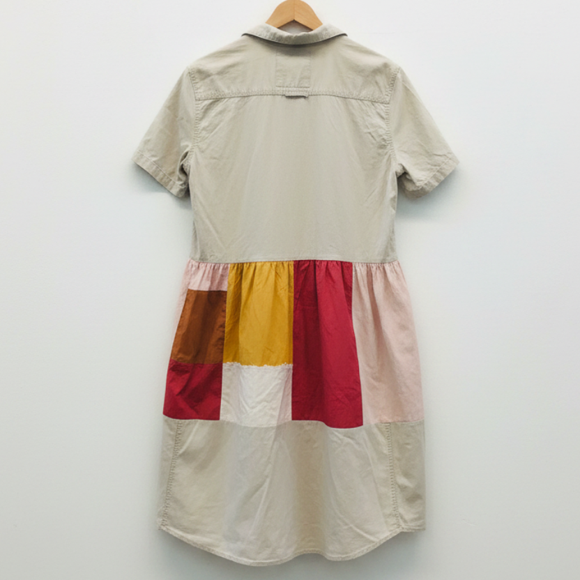 A short-sleeve patchwork shirt dress hanging on a wooden hanger against a white painted brick wall. The dress features a light beige upper body and skirt, with a gathered waistband and colorful patchwork panels in mustard yellow, red, and brown on the back.