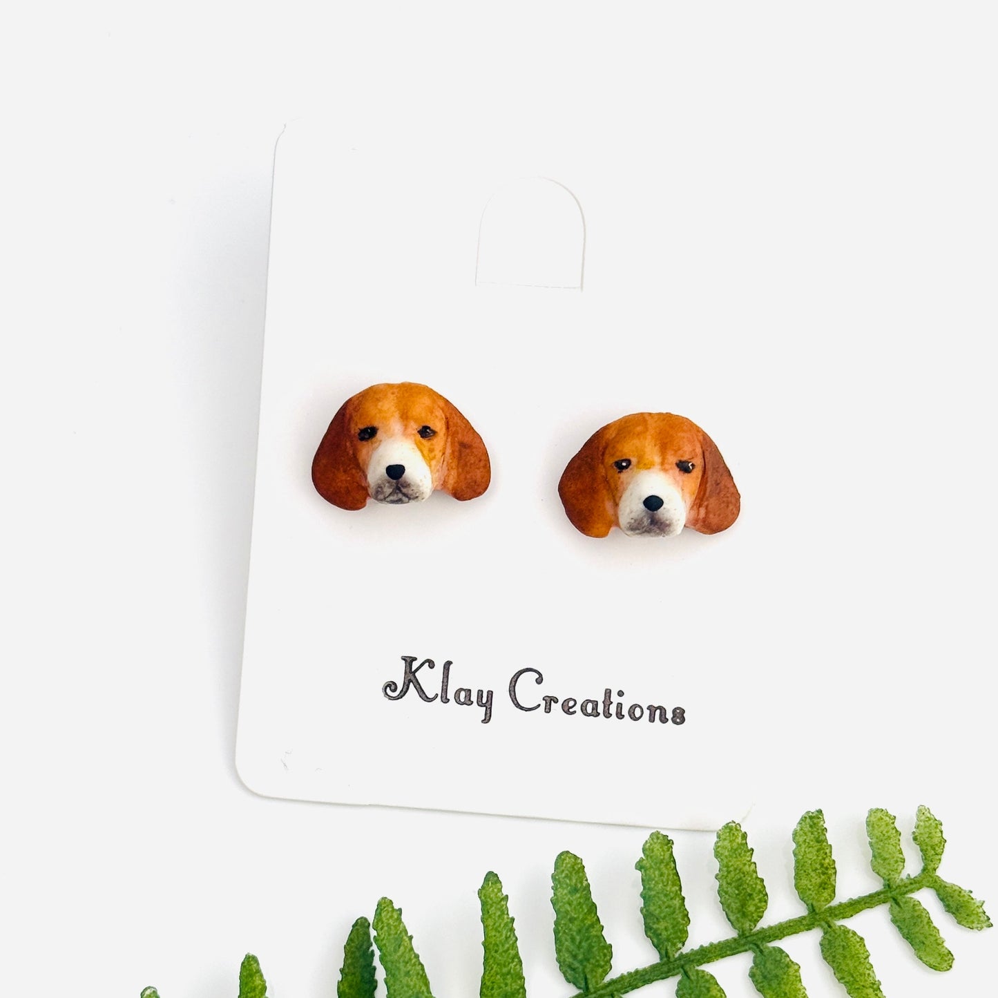A pair of stud earrings shaped like beagle dog faces, displayed on a white backing card with the brand name 'Klay Creations' printed below. A green fern leaf is placed diagonally in the foreground on a clean white background.