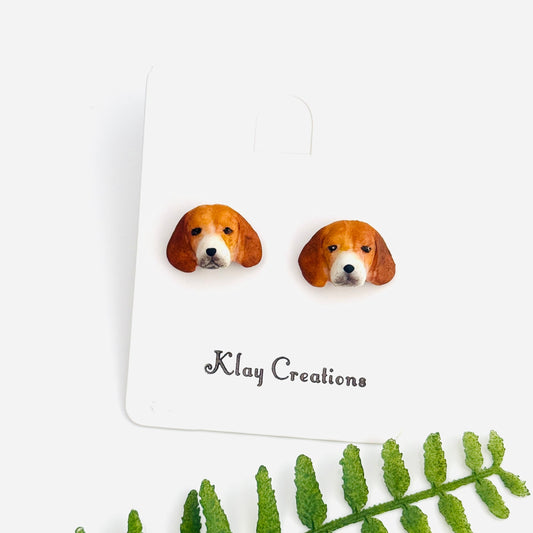A pair of stud earrings shaped like beagle dog faces, displayed on a white backing card with the brand name 'Klay Creations' printed below. A green fern leaf is placed diagonally in the foreground on a clean white background.