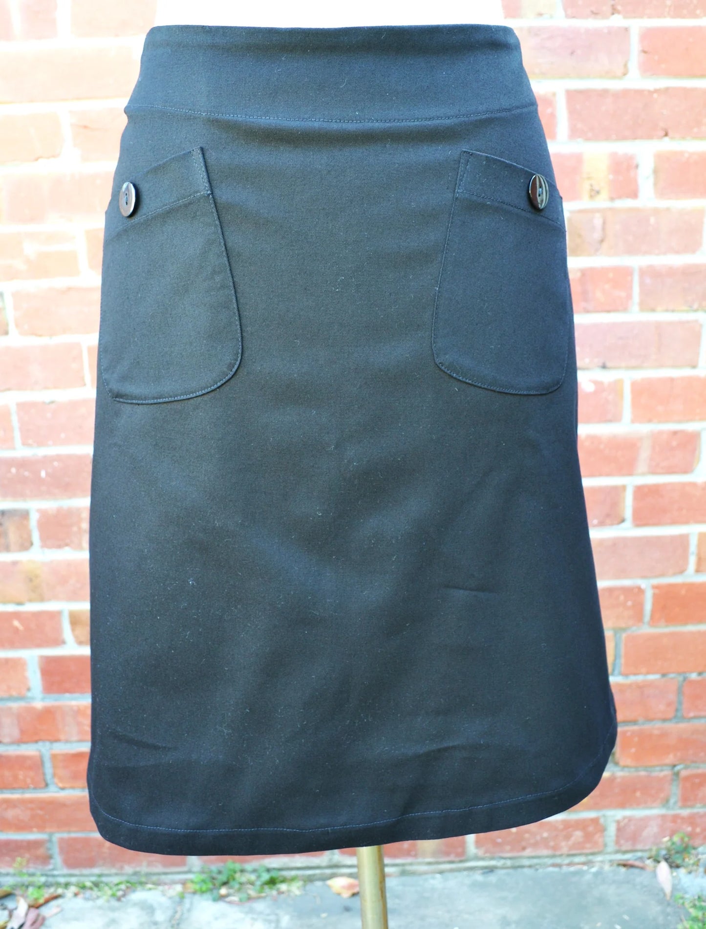 Black skirt with two front pockets on a stand against a brick wall.