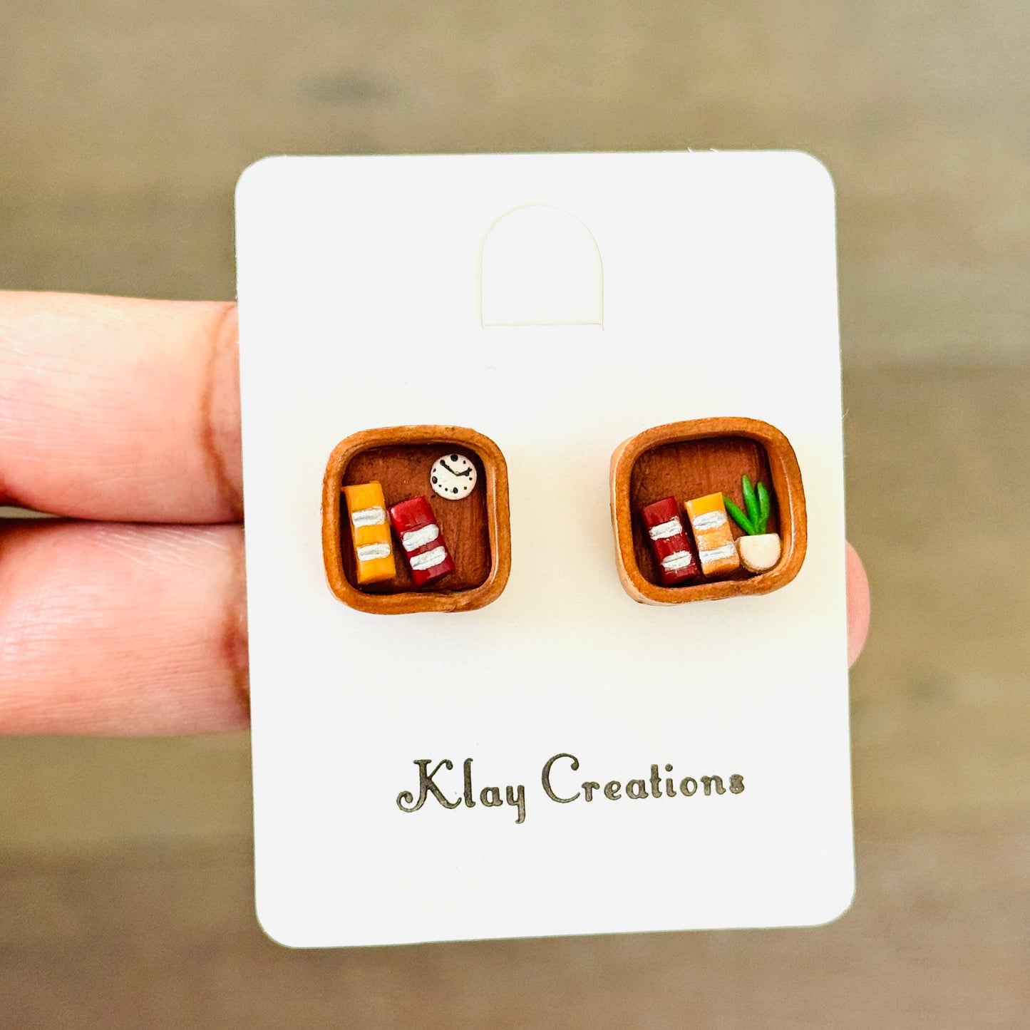 A pair of miniature food-themed stud earrings displayed on a white backing card held by a hand. The earrings are set in a warm brown frame and depict tiny illustrations of sushi, tacos, and a sandwich.