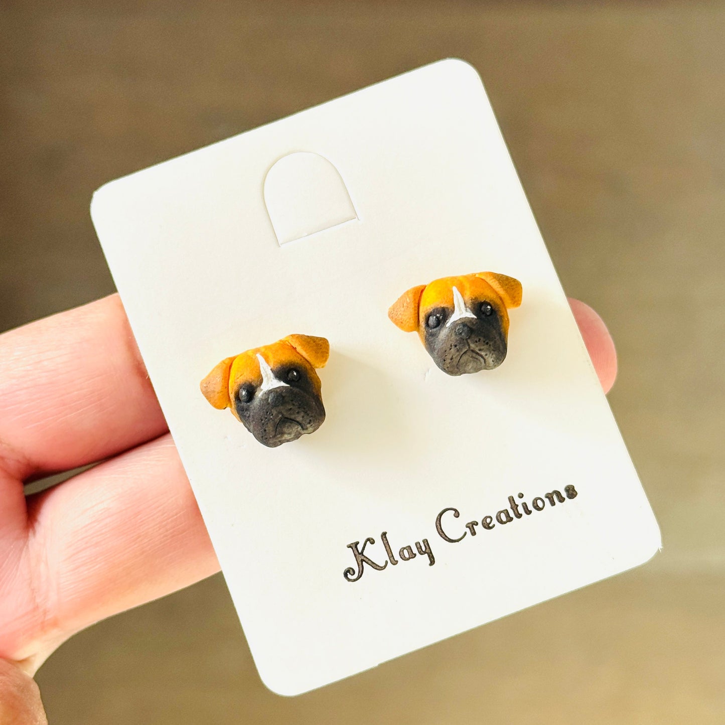 A hand holding a white display card featuring a pair of stud earrings shaped like dog faces. The earrings have tan and white fur with black eyes and noses.