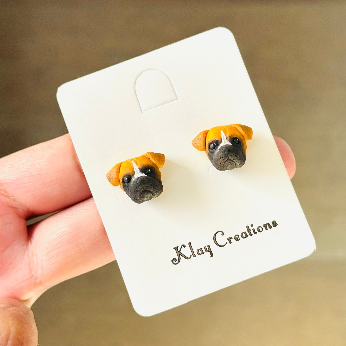 A hand holding a white display card featuring a pair of stud earrings shaped like dog faces. The earrings have tan and white fur with black eyes and noses.