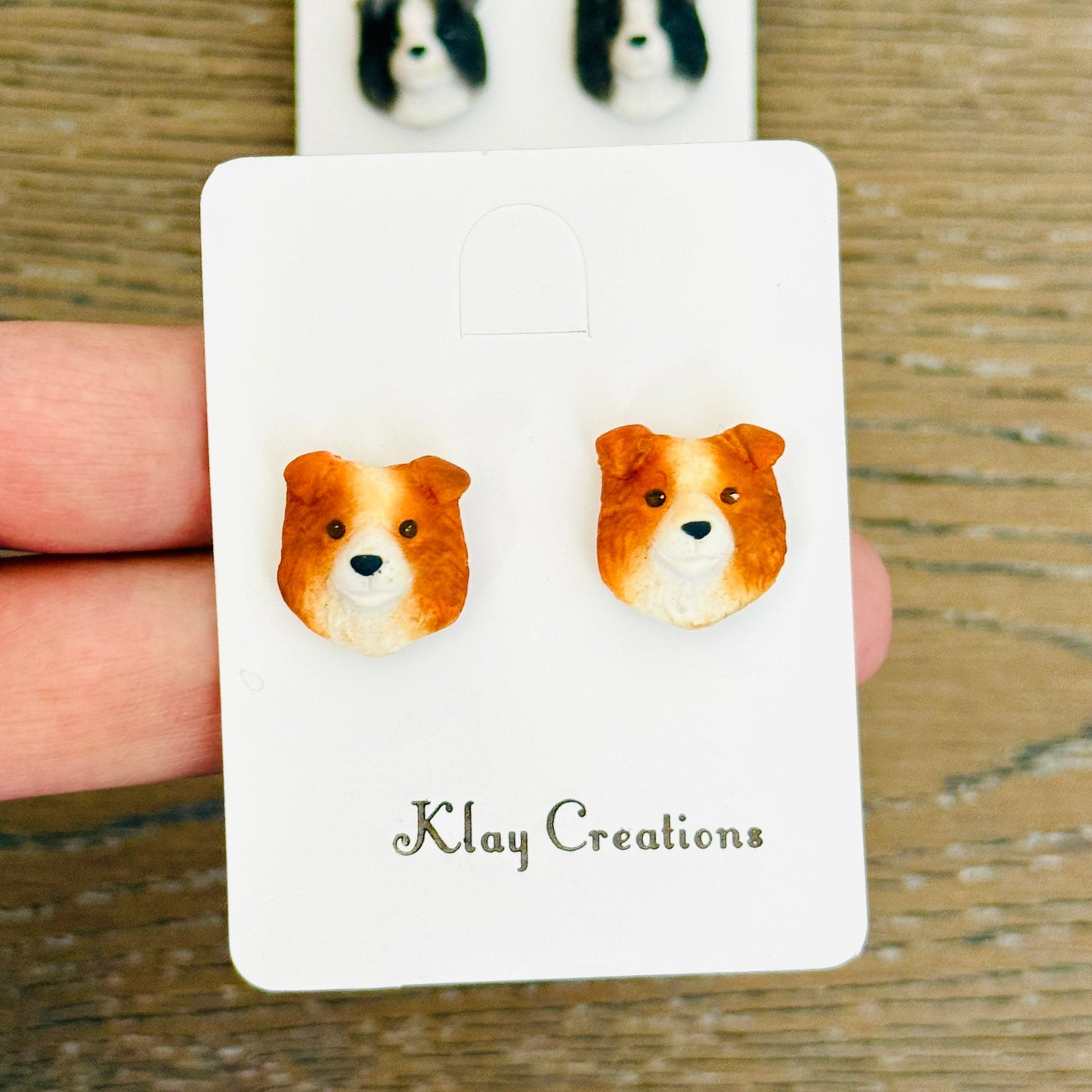 A pair of small dog-face stud earrings displayed on a white backing card held by a hand over a wooden surface. The front card shows two orange-and-white dog stud earrings, and a second pair of black-and-white dog stud earrings is visible behind on a larger backing card.
