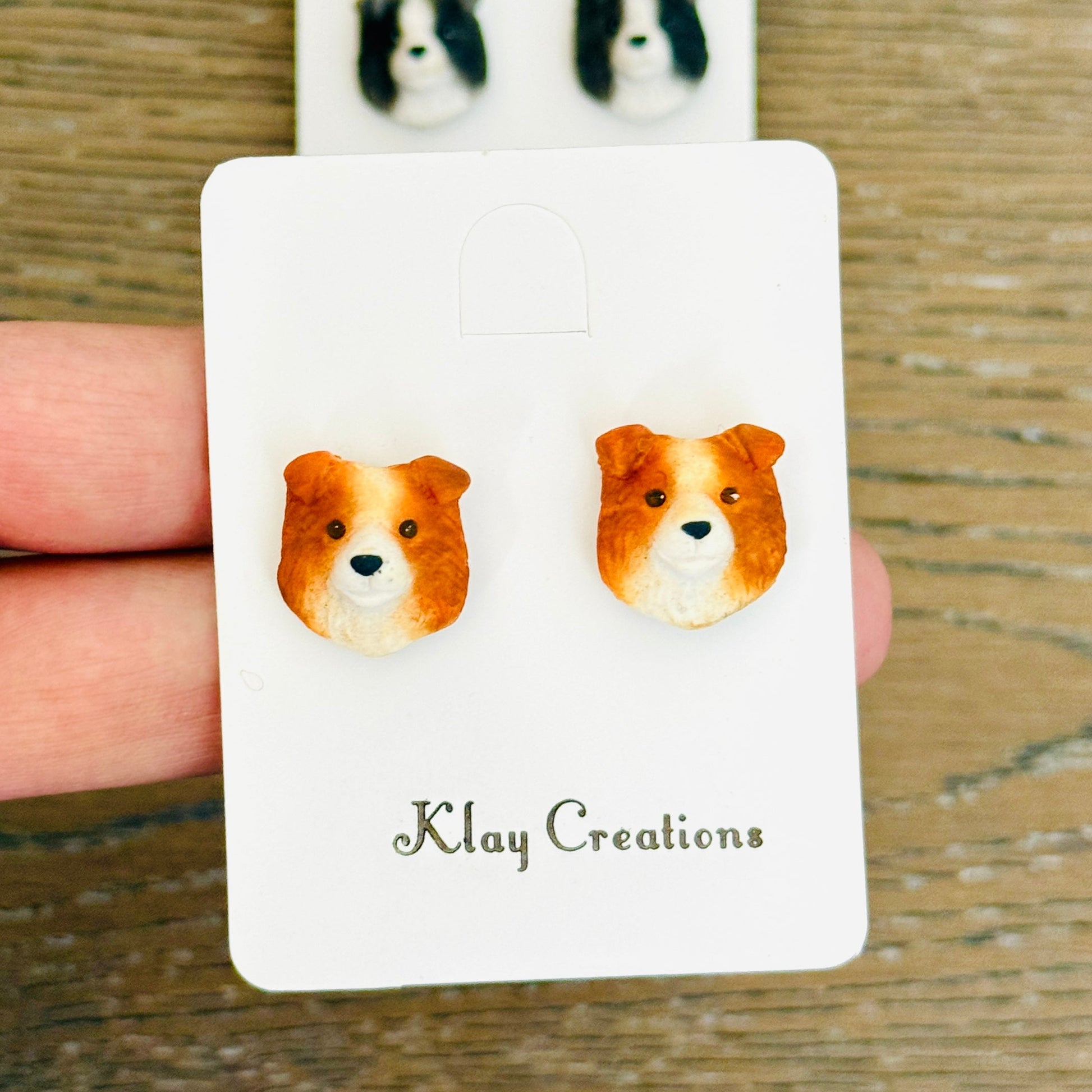 A pair of small dog-face stud earrings displayed on a white backing card held by a hand over a wooden surface. The front card shows two orange-and-white dog stud earrings, and a second pair of black-and-white dog stud earrings is visible behind on a larger backing card.