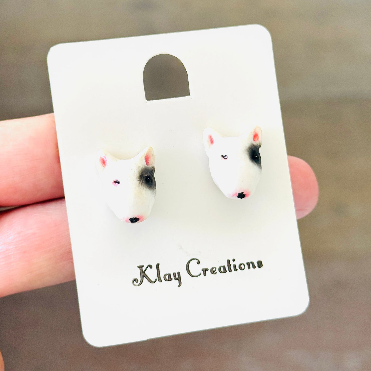 A pair of small stud earrings shaped like dog faces, mounted on a white display card held by a hand. The card is branded 'Klay Creations' and is photographed against a blurred brown background.
