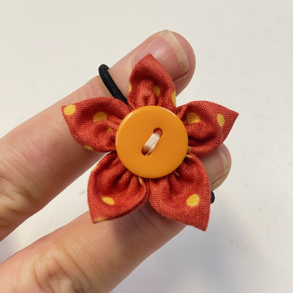 Hand holding a red fabric flower hairtie with a yellow button center against a white background