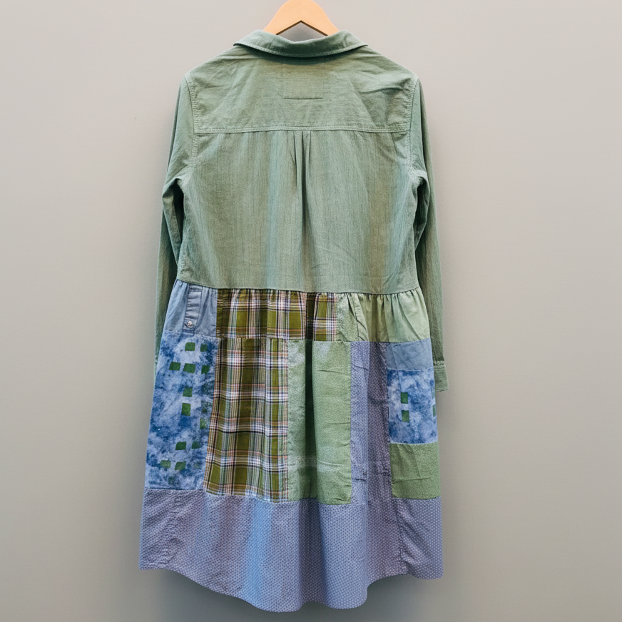 A long-sleeve patchwork dress hanging on a wooden hanger against a plain light gray wall. The back view shows a sage green upper section, a gathered waistband, and patchwork lower panels in blue floral, green-and-tan plaid, and light blue gingham.