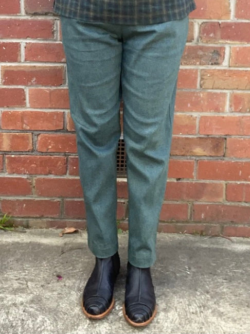 Person wearing green pants and dark shoes standing against a brick wall.