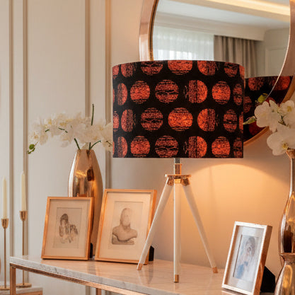 A mid-century modern tripod table lamp with a black fabric shade featuring a pattern of orange-red textured circles, a copper-finished central collar, and three white metal legs with copper tips.