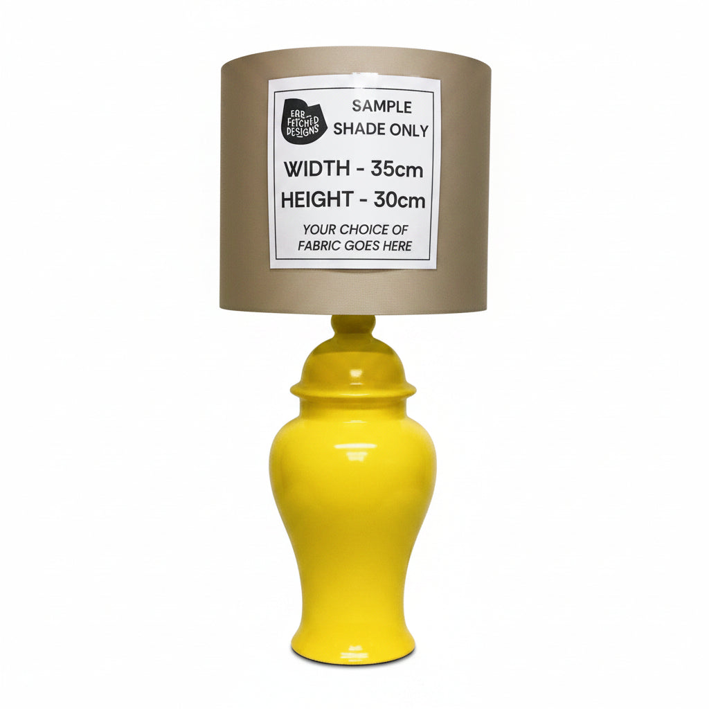 A table lamp with a glossy yellow ceramic vase-style base and a brown fabric shade, shown against an off-white wall. A white paper label on the shade reads 'SAMPLE SHADE ONLY' with dimensions and fabric placeholder text.