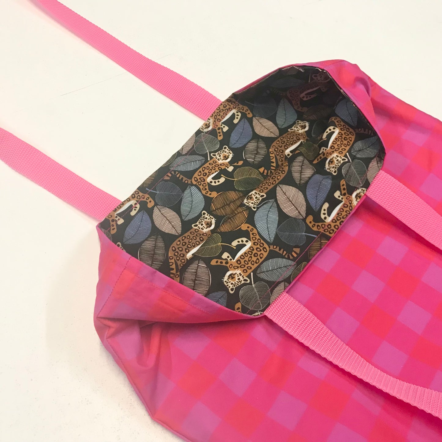 A pink tote bag with a pink and magenta checkered exterior, bright pink handles, and a dark patterned interior featuring a leopard and leaf print.