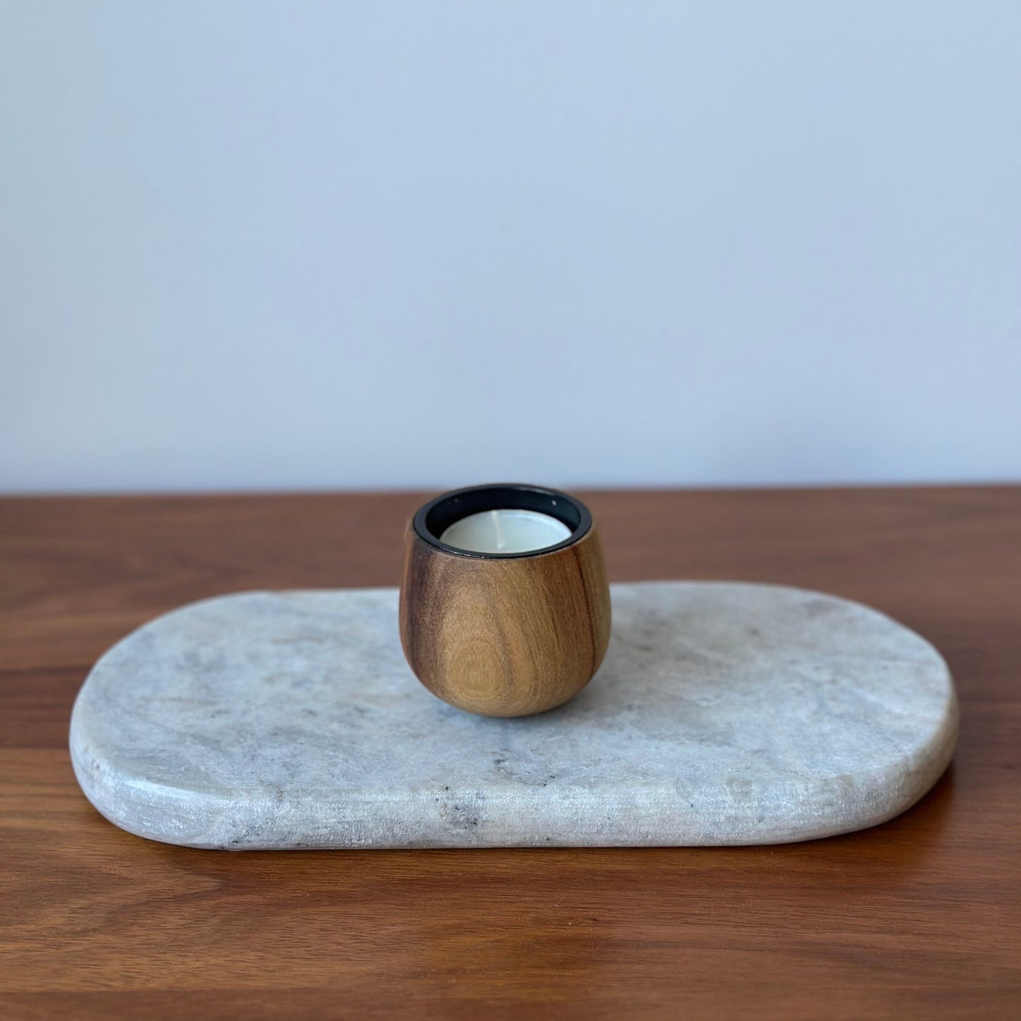A small wooden candle holder with a black rim and white wax inside, displayed on an oval light gray stone tray over a wooden surface against a light gray wall.
