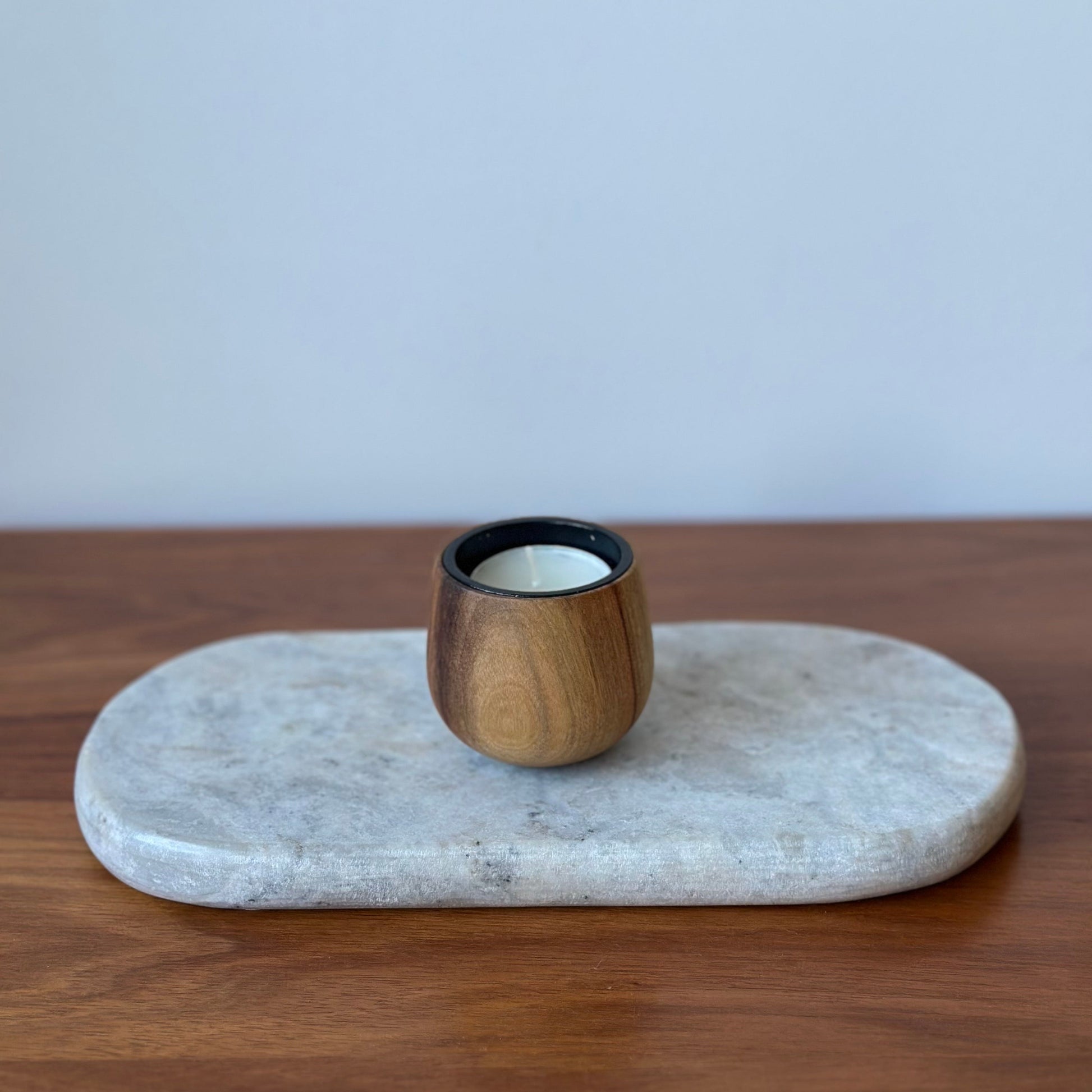A small wooden candle holder with a black rim and white wax inside, displayed on an oval light gray stone tray over a wooden surface against a light gray wall.