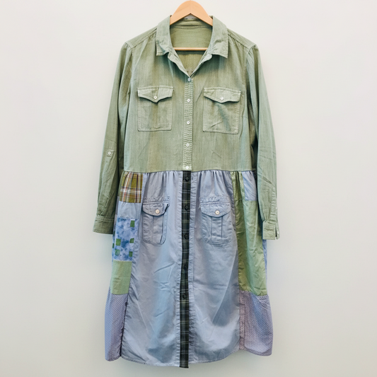 A patchwork button-up shirt dress hanging on a wooden hanger against a white-painted brick wall. The dress features a sage green upper section with two chest pockets, a light blue denim-like lower panel with two front flap pockets, and a central black plaid stripe running down the front.