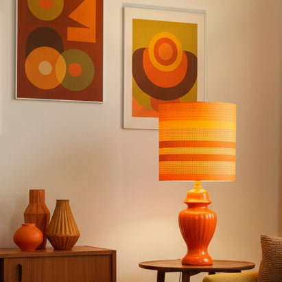 A table lamp with a glossy orange fluted glass base and a matching orange fabric drum shade featuring horizontal stripes in orange, yellow, and brown.